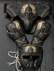 A coordinated set of black protective gear with gold bat-themed graphics, including a headguard, boxing gloves, and a waist guard, arranged on a dark wooden surface.