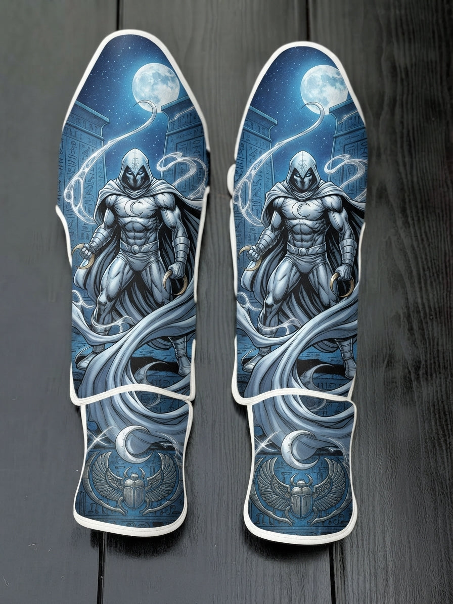 Custom Moon Knight superhero design with white hooded cloak, crescent darts, full moon, Egyptian hieroglyphics, and winged scarab emblem soccer shin guards displayed on a dark surface.