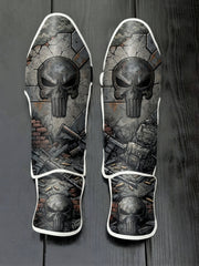 Custom Punisher superhero design with weathered metallic skull logo, tactical military gear, scattered ammunition, and distressed concrete wall soccer shin guards displayed on a dark surface.