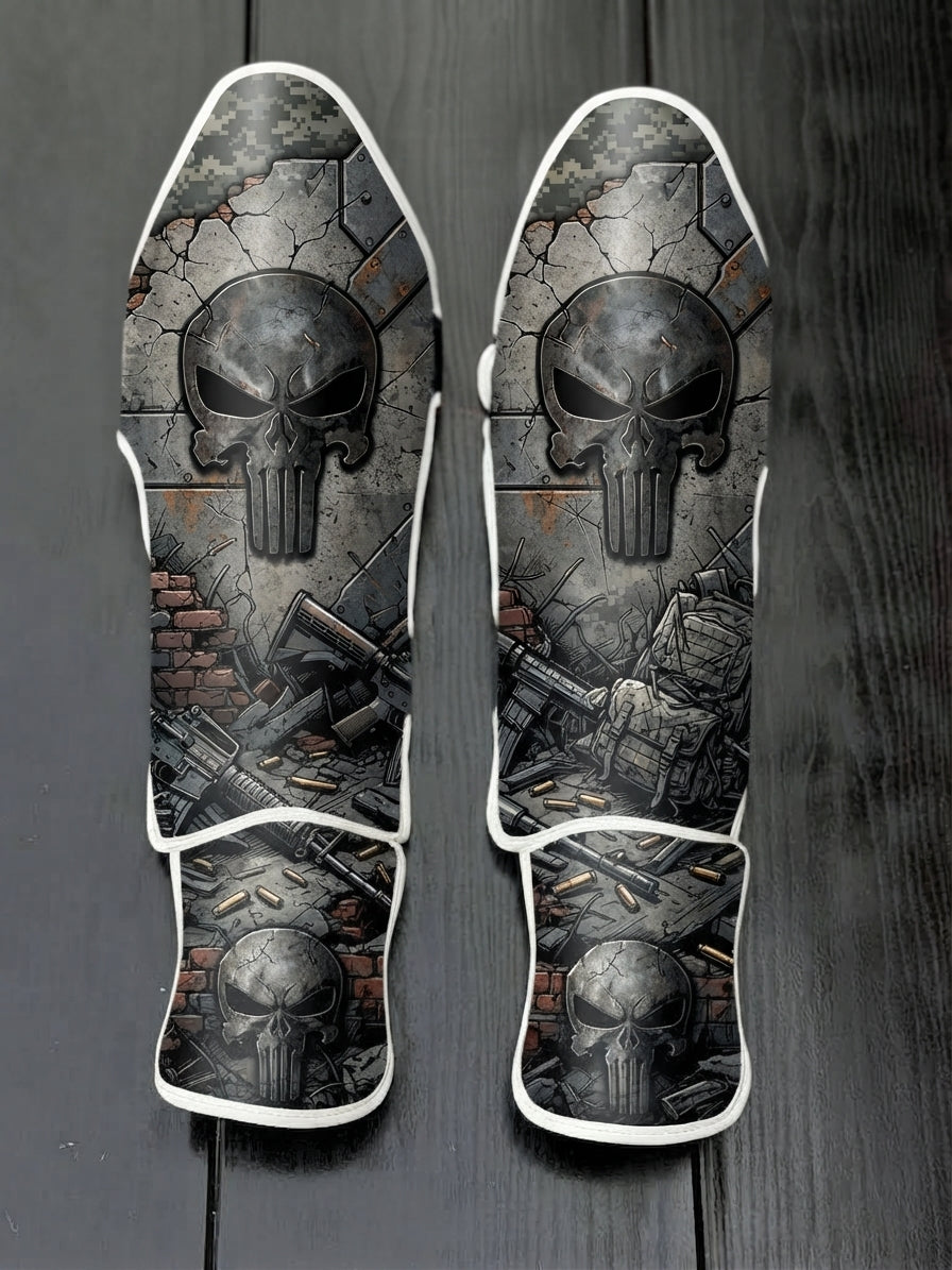 Custom Punisher superhero design with weathered metallic skull logo, tactical military gear, scattered ammunition, and distressed concrete wall soccer shin guards displayed on a dark surface.