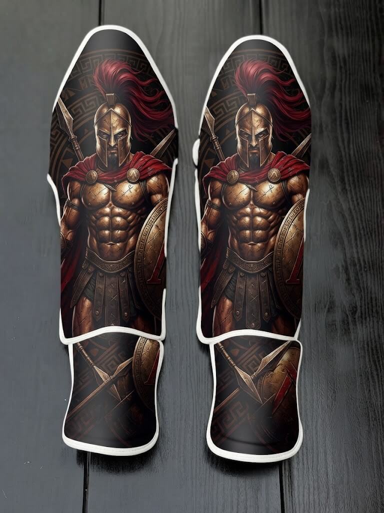 Custom Spartan warrior design with bronze Corinthian helmet, red horsehair plume, cape, muscular physique, spear, shield, and Greek geometric border background soccer shin guards displayed on a dark surface.