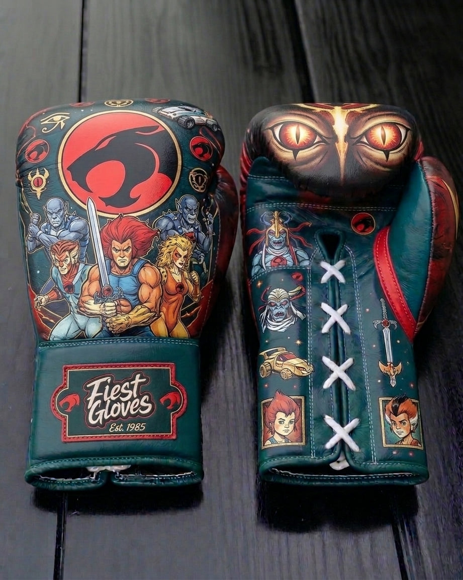 A close-up photograph of a pair of detailed, dark teal green leather boxing gloves featuring classic ThunderCats artwork. The left glove shows Lion-O and other heroes under the red cat logo, and the right glove displays giant menacing Mumm-Ra eyes. Both cuffs bear the label "Fierst Gloves Est. 1985.